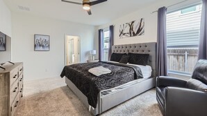 Quarto com cama, ventilador de teto, janela com cortinas, cômoda e uma cadeira.