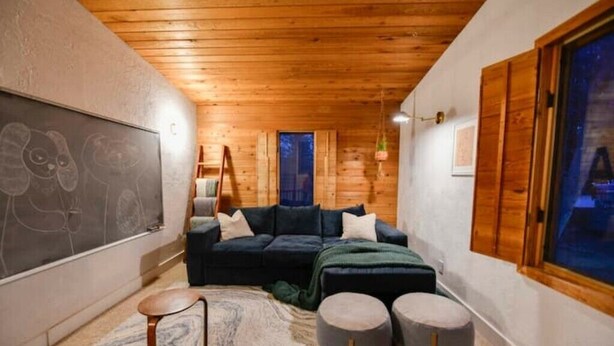 A cozy room with a dark blue sofa, a small round table, and two ottomans. A chalkboard with drawings is on the wall, and there's a window with wooden shutters. - Cozy and Artistic Space with a library and a Floating Net (Idaho Falls)
