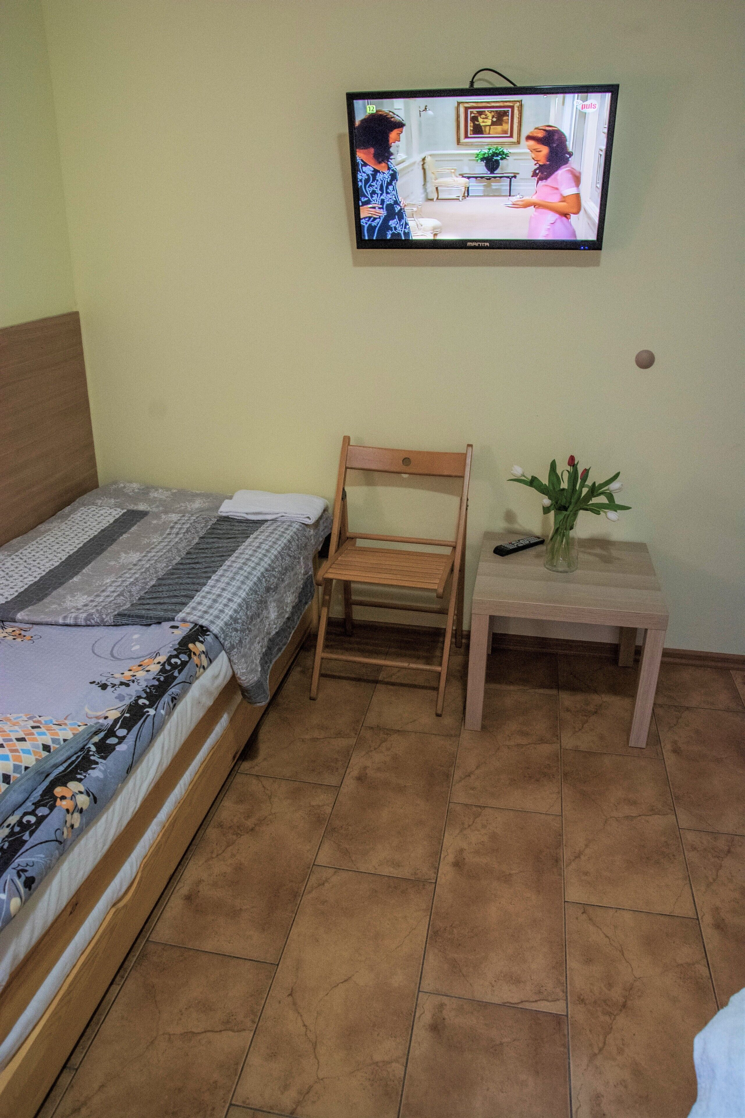 A bedroom with a bed, a wooden chair, a small table with a vase of flowers, and a television mounted on the wall.