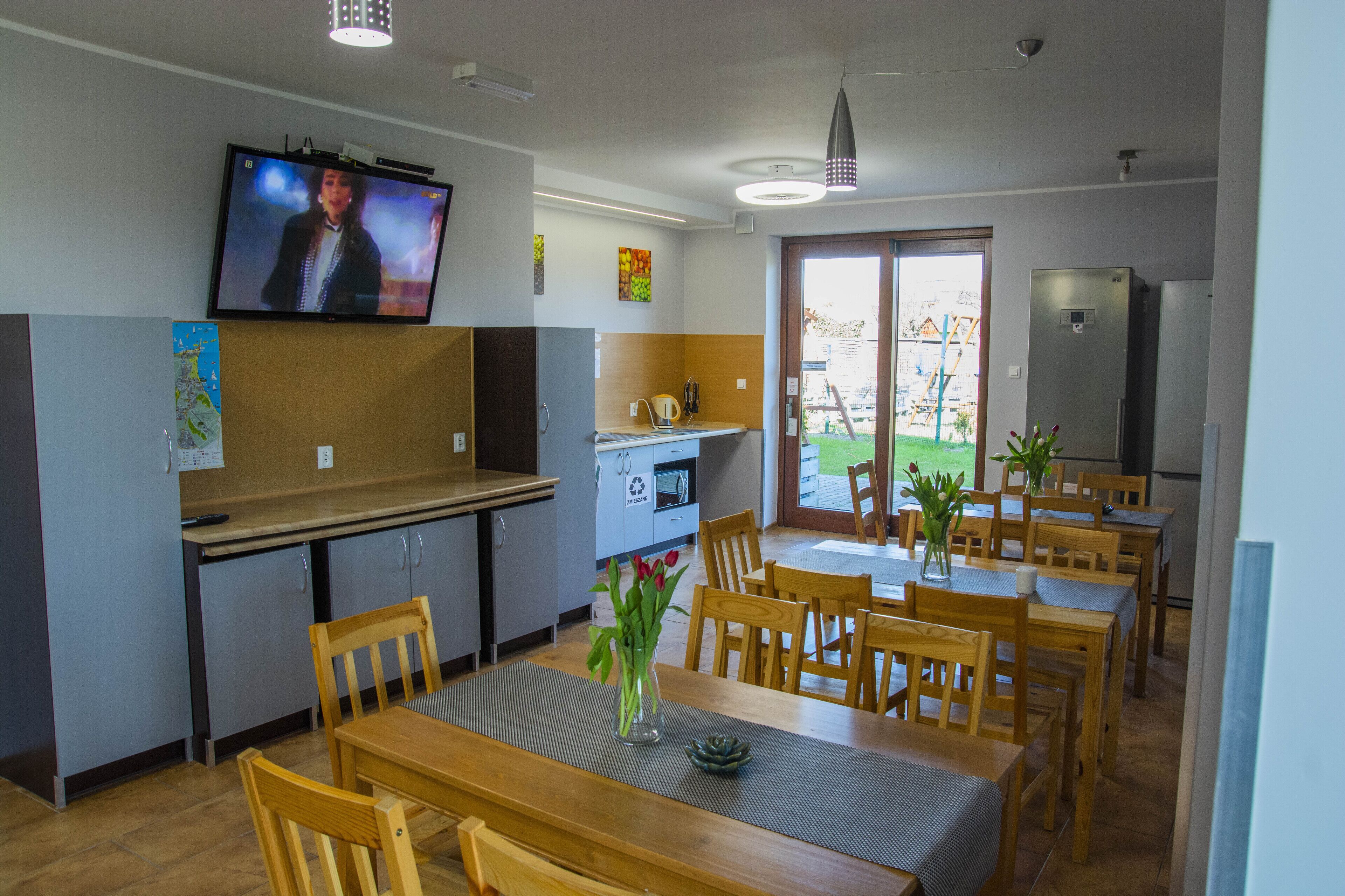 A dining area with wooden tables and chairs, a television mounted on the wall, and a refrigerator in the background.