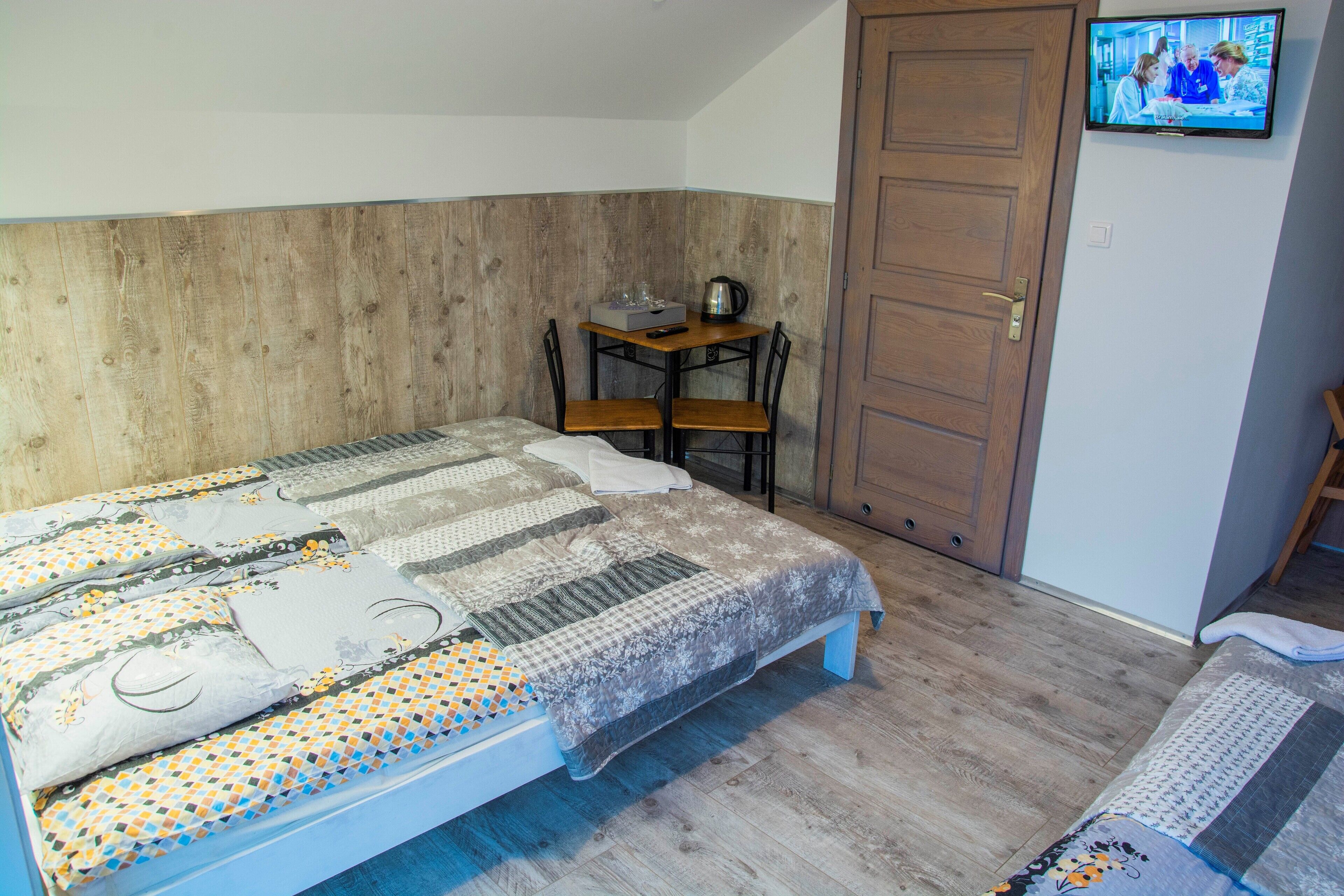 A bedroom with a bed, a wooden headboard, a small table with a kettle, and a TV on the wall.