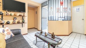 A living room with a sofa, a coffee table with decorative items, a flat-screen TV mounted above a shelf with various objects, and a door with a sign indicating social distancing measures. - Kurmi House Lima Airport (Callao)