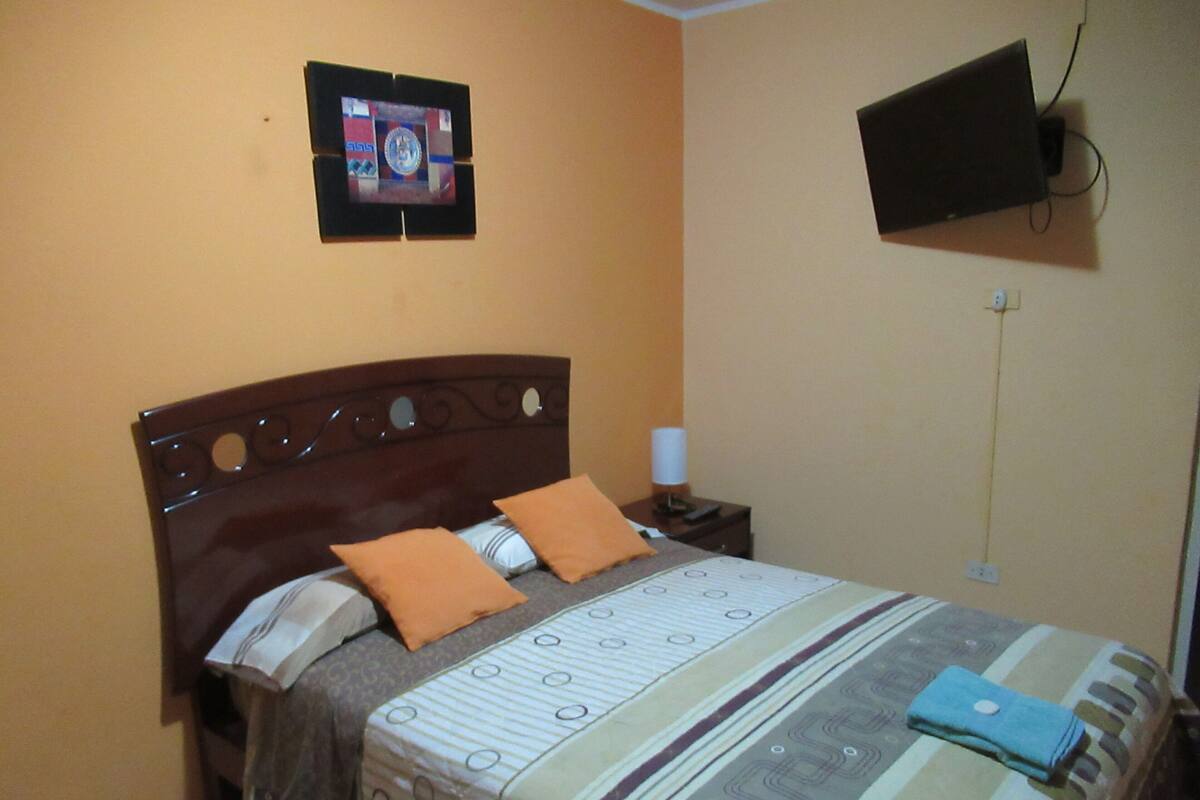 A hotel room with a bed, a TV mounted on the wall, and a bedside table with a lamp.