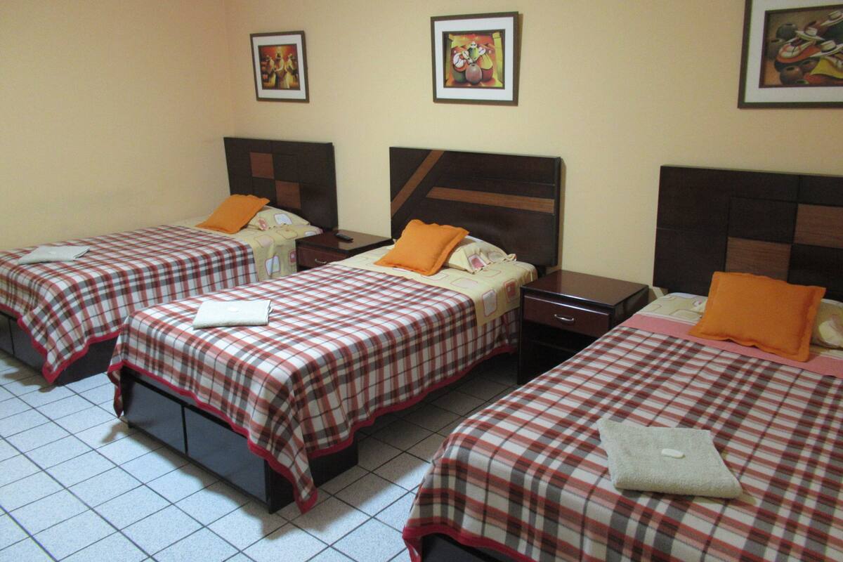 A hotel room with three beds, each with a checkered bedspread and orange pillows. There are framed pictures on the wall and a tiled floor.