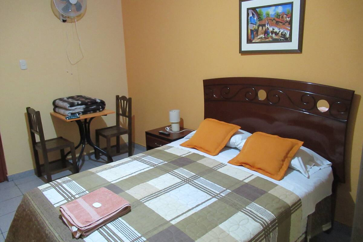 A neatly made bed with an orange pillow, a bedside table with a lamp, a chair, and a desk with a fan.