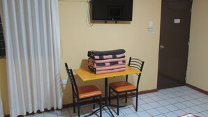 A small room with a bed, a TV mounted on the wall, a table with chairs, and a door. - Kurmi House Lima Airport (Callao)