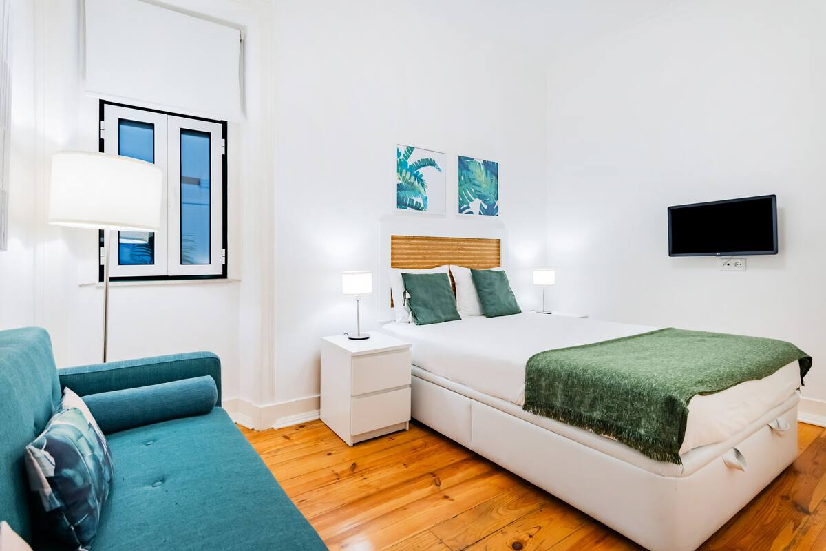 Ein Schlafzimmer mit Bett, Nachttisch, Fernseher, Fenster und einer Couch.