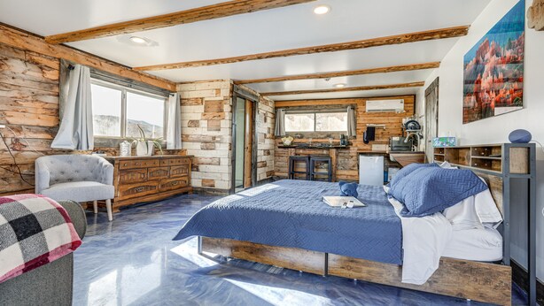 A bedroom with a wooden bed frame, a dresser, a chair, and a view of a kitchen area. - Cozy Parowan Studio: 12 Mi to Brian Head Resort! (Parowan)