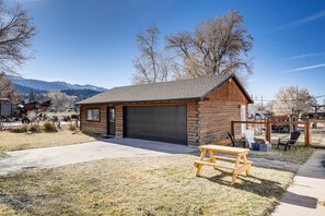 A wooden cabin with a garage, a picnic table, and a fenced area with chairs.