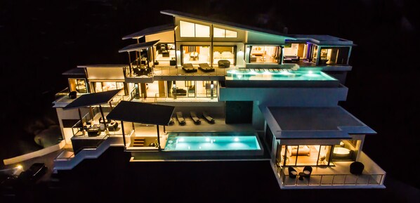 A multi-story modern house with a swimming pool, outdoor seating, and a well-lit interior.