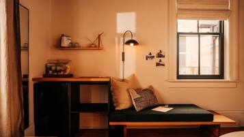 A cozy room with a wooden desk, a green cushioned bench, a floor lamp, and a window with blinds.