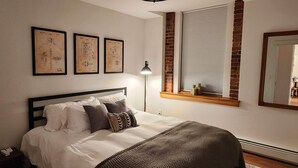 Une chambre à coucher comprenant un lit, une table de chevet, une lampe, une fenêtre avec des stores et trois dessins encadrés accrochés au mur.