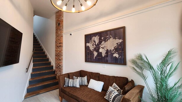 Un escalier avec un mur en briques, un canapé marron avec des coussins à motifs, une œuvre d’art représentant une carte du monde et une grande plante en pot.