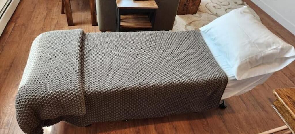 Um quarto pequeno e acolhedor com uma cama de solteiro, uma mesinha de cabeceira e um travesseiro branco.