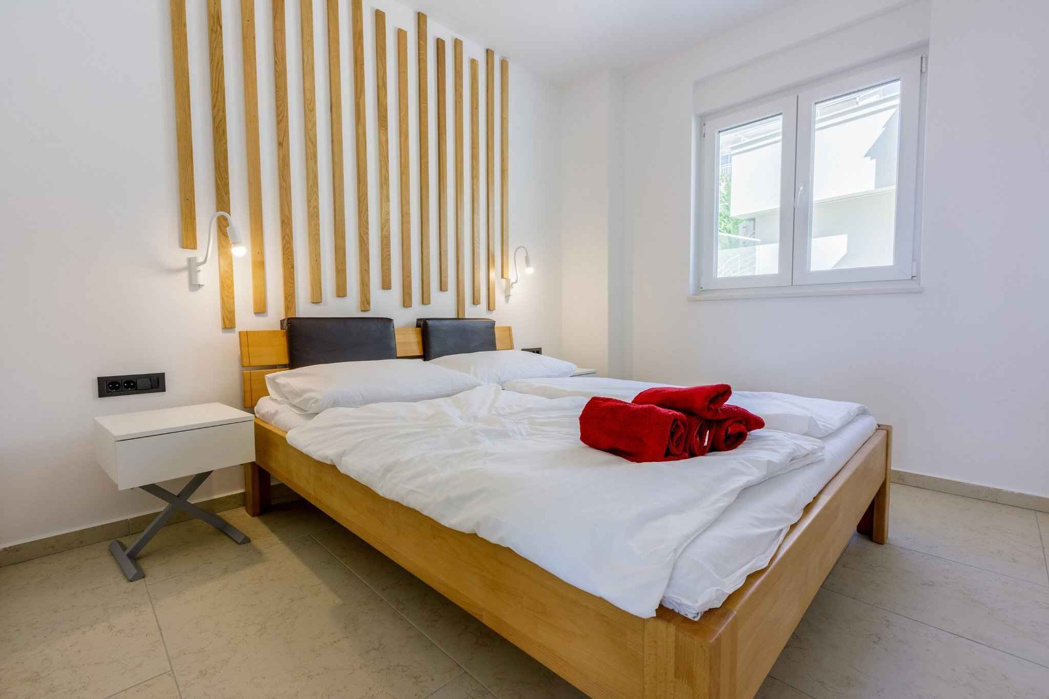 A modern bedroom with a wooden bed, white bedding, a bedside table, a wall-mounted light fixture, and a window with blinds.