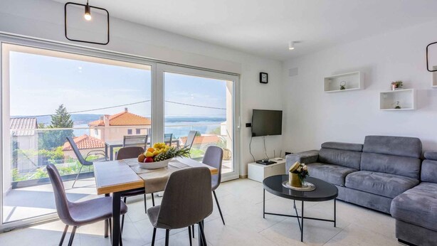 A modern living room with a grey sofa, a dining table with chairs, a TV on a stand, and a balcony with a view of the sea and buildings. - Holiday apartment with shared-use pool (Crikvenica)
