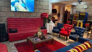 Una sala de estar con un sofá rojo, sillones azules y una mesa de centro. Un televisor está montado en la pared, y hay una chimenea con un reloj encima.