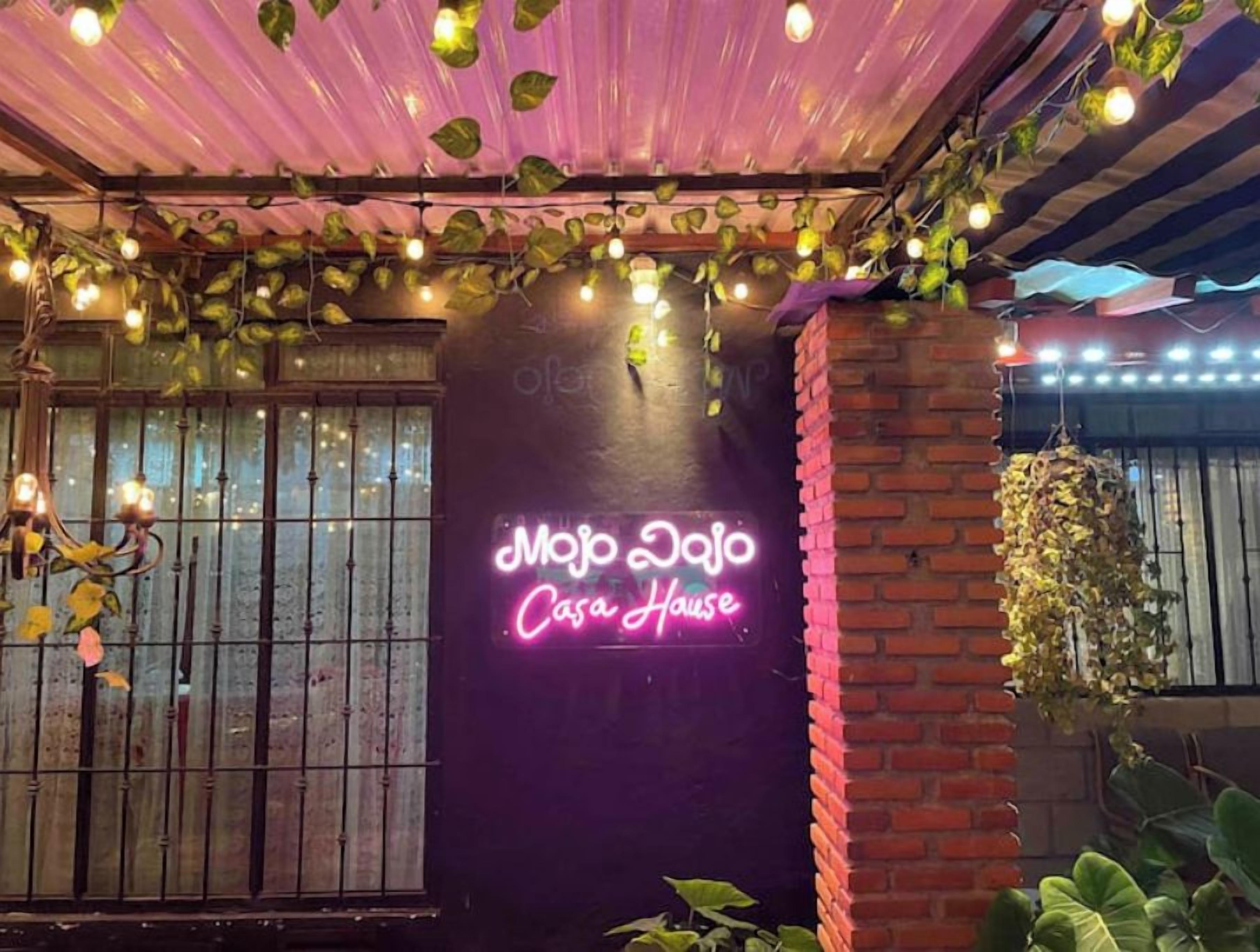 A neon sign reading "Mojo Dojo Casa House" with a brick wall and hanging plants.