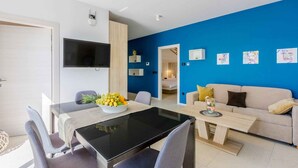 A modern living room with a blue accent wall, a sofa, a dining table with chairs, and a flat-screen TV mounted on the wall. - Holiday apartment with shared-use pool (Crikvenica)