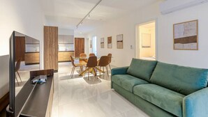 Ein modernes Wohnzimmer mit einem grünen Sofa, einem Essbereich mit einem Glastisch und Stühlen, und einer Küche im Hintergrund.