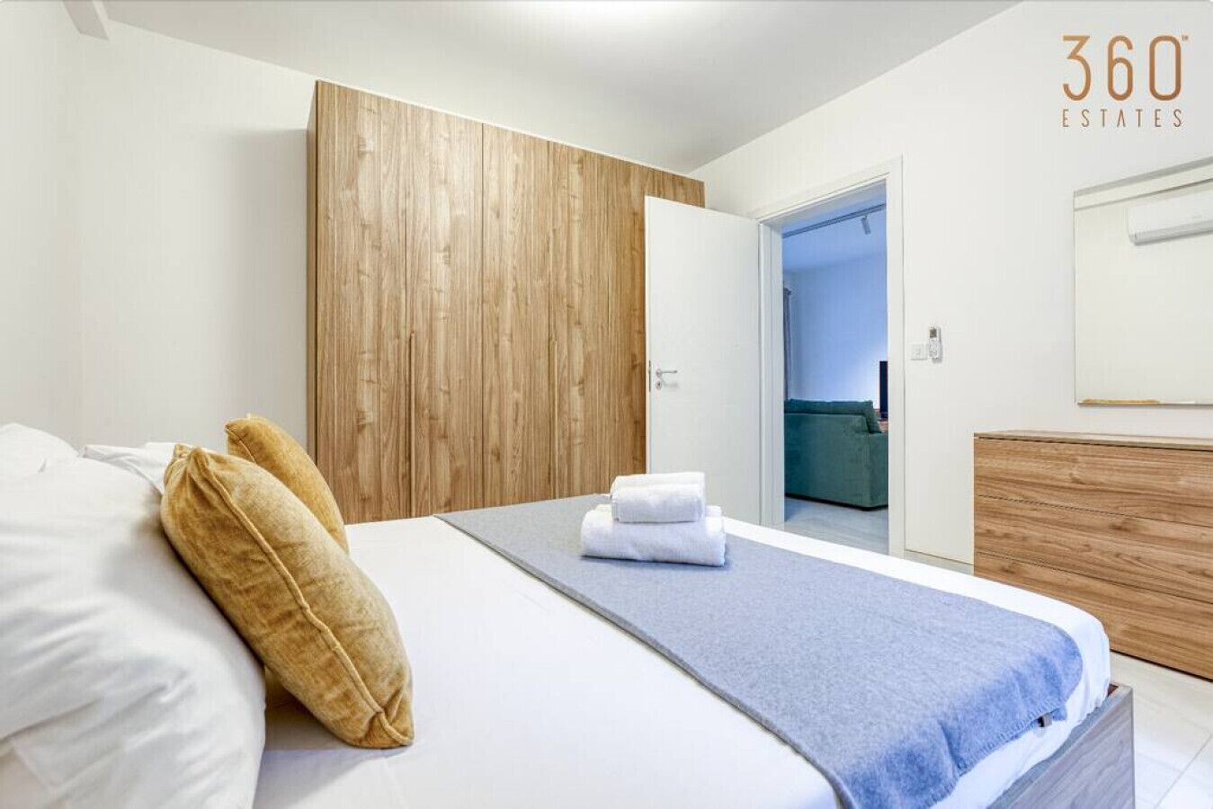 Een moderne slaapkamer met een houten garderobe, een bed met witte lakens en een blauwe deken, en een zichtbare badkamer op de achtergrond.