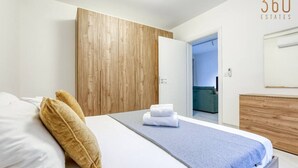Ein modernes Schlafzimmer mit einem Holzschrank, einem Bett mit weißen Bettwäsche und einer blauen Decke, sowie ein sichtbares Badezimmer im Hintergrund.