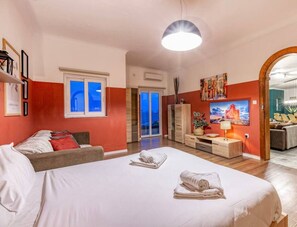 Une chambre spacieuse avec un grand lit, un canapé, une télévision et un coin repas visible à travers une porte ouverte.