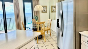 A modern kitchen with a stainless steel refrigerator, a dining area with a glass table and wooden chairs, and a view of the ocean through large windows. - Stunning Views from this 15th floor one bedroom condo! (Orange)