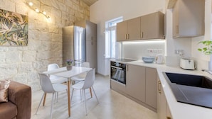 A modern kitchen with a stone wall, stainless steel appliances, and a dining area with a white table and chairs. - Charming + Modern 1BR House in Historic Zebbug (Ħaż-Żebbuġ)