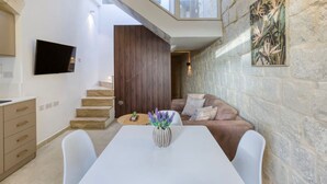 A modern living space with a dining area, a sofa, a TV mounted on the wall, and a staircase leading to another level. - Charming + Modern 1BR House in Historic Zebbug (Ħaż-Żebbuġ)