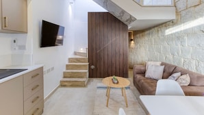 A modern kitchen with a built-in oven, a dining area with a white table and chairs, a brown sofa with cushions, and a stone wall. - Charming + Modern 1BR House in Historic Zebbug (Ħaż-Żebbuġ)