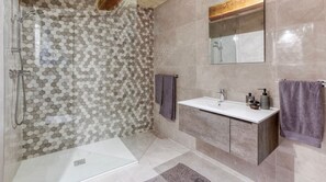 Un baño moderno con pared de ducha revestida de azulejos hexagonales, cabina de ducha con mampara de vidrio, lavabo blanco con mueble de madera y un espejo grande sobre el lavabo.