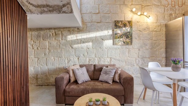 A cozy living room with a stone wall, a brown sofa, a wooden coffee table with plants, and a white dining area. - Charming + Modern 1BR House in Historic Zebbug (Ħaż-Żebbuġ)