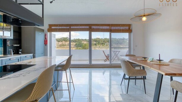 A modern kitchen with a large island, dining area with a long table and chairs, and a view through large windows. - A lovely, central 2BR penthouse with large outdoor (L-Iklin)