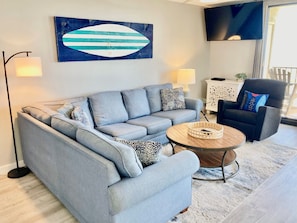 A modern living room with a grey sofa, a round coffee table, and a blue and white abstract painting on the wall. - NEWLY RENOVATED 3 Bedroom CORNER unit on the 4th Floor With Gulf Views! (Orange Beach)