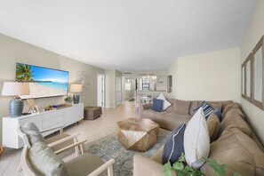 A spacious living room with a beige sofa, a coffee table, and a TV displaying a beach scene. There is a dining area in the background with a table and chairs.