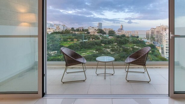 A balcony with two wicker chairs and a small table, overlooking a cityscape with buildings and greenery. - A fully equipped 3BR home with private balcony (San Ġiljan)