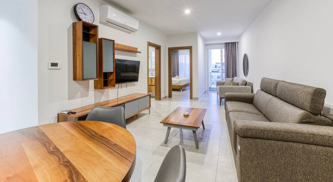 A modern living room with a wooden dining table, a sofa, a TV mounted on the wall, and a bed in the background.