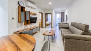 Ein modernes Wohnzimmer mit einem Holzesstisch, einer Couch, einem an der Wand montierten Fernseher und einem Bett im Hintergrund.