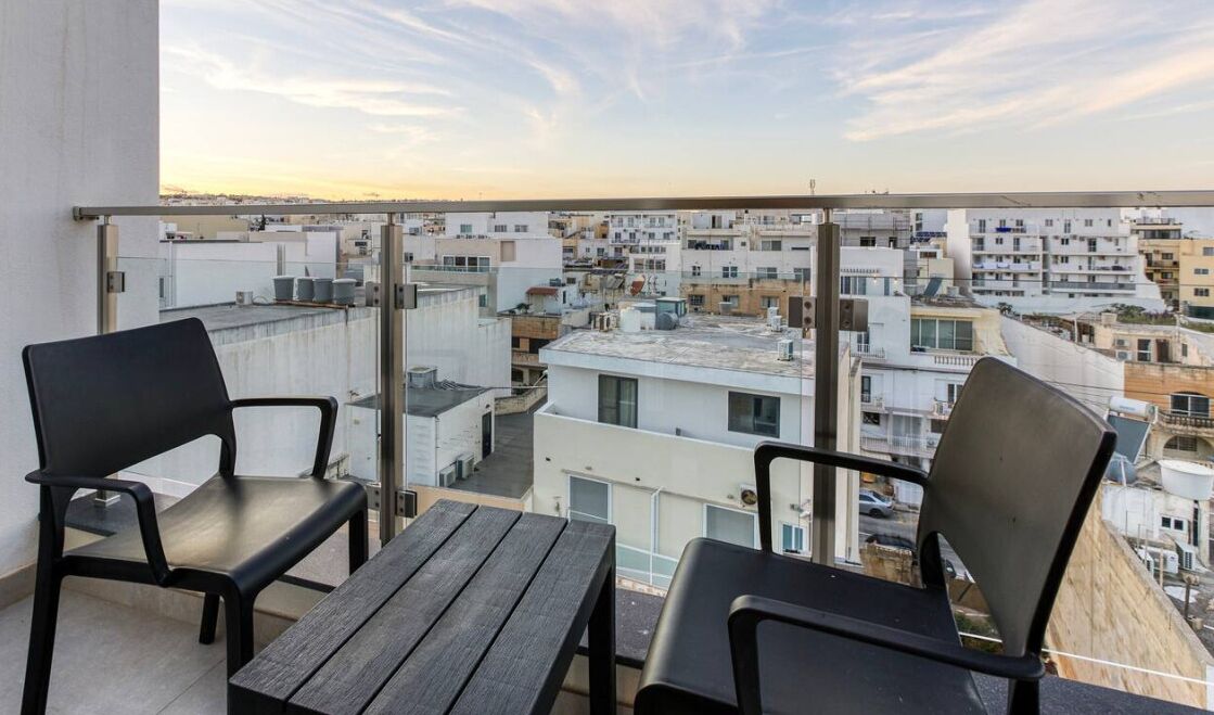 A balcony with two black chairs and a wooden table, overlooking a densely packed urban area with various buildings.