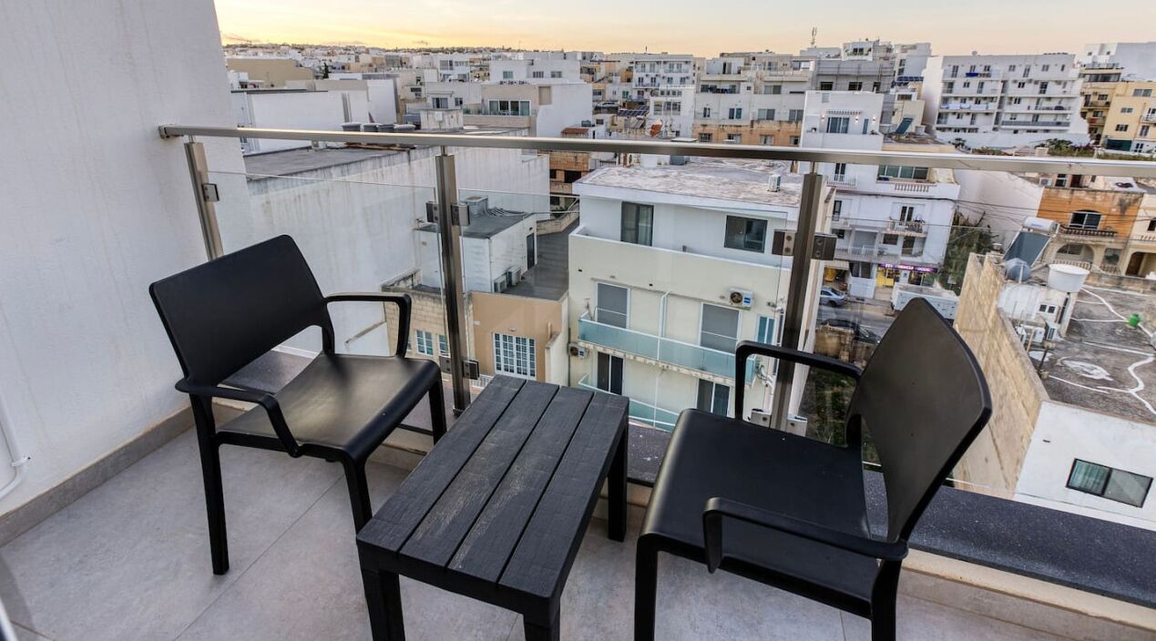 Ein Balkon mit zwei schwarzen Stühlen und einem kleinen Tisch, der auf einen dicht bebauten Stadtbereich mit verschiedenen Gebäuden blickt.