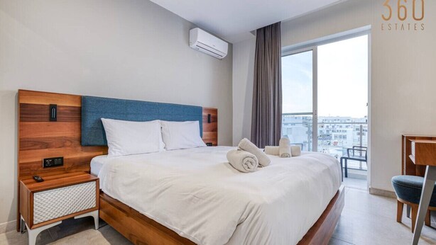 Ein modernes Hotelzimmer mit einem großen Bett, einem Nachttisch, einem Sessel und Blick auf die Stadt durch große Fenster.