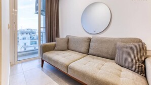 Ein modernes Wohnzimmer mit einer Couch, zwei gemusterten Kissen, einem runden Spiegel und einer Schiebetür zum Balkon.