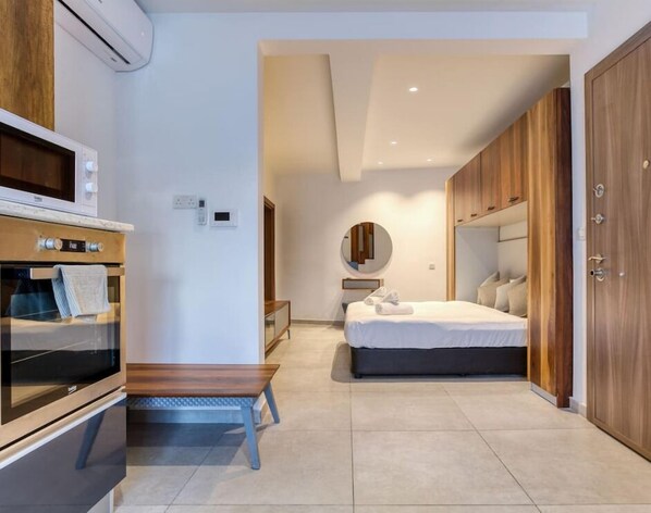Une chambre d’hôtel moderne avec une armoire en bois, un lit doté d’une tête de lit sombre, un miroir rond et un banc intégré.
