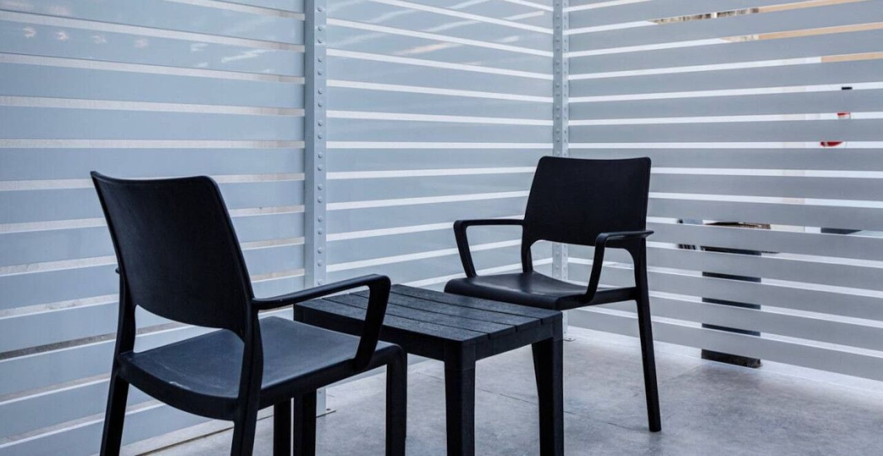 Un espacio moderno al aire libre con dos sillas negras y una mesita pequeña, frente a una pared metálica con listones horizontales.