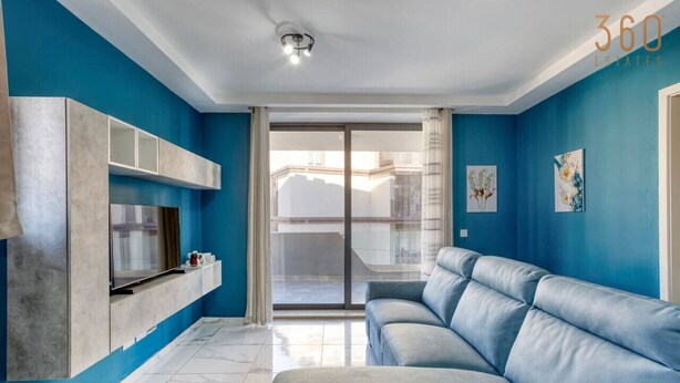 A modern living room with a blue wall, a grey sofa, a TV mounted on a grey cabinet, and a sliding glass door to the balcony. - Relaxing 2BR home with Private Balcony in Mellieha (Il-Mellieħa)