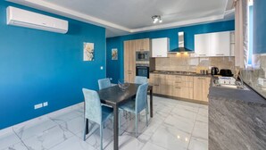 A modern kitchen with blue walls, a dining area with a table and chairs, and a built-in oven. - Relaxing 2BR home with Private Balcony in Mellieha (Il-Mellieħa)