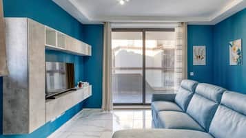 Una sala de estar moderna con una pared azul, un sofá gris, un televisor montado en un mueble gris y un ventanal corredera al balcón.