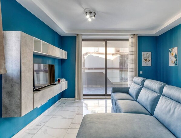 Un salon moderne avec un mur bleu, un canapé gris, un téléviseur fixé sur un meuble gris et une porte-fenêtre coulissante donnant sur le balcon.