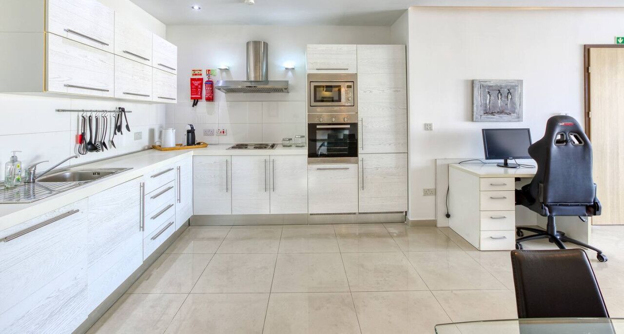 A modern kitchen with white cabinetry, a built-in oven, and a small dining area with a desk and chair.